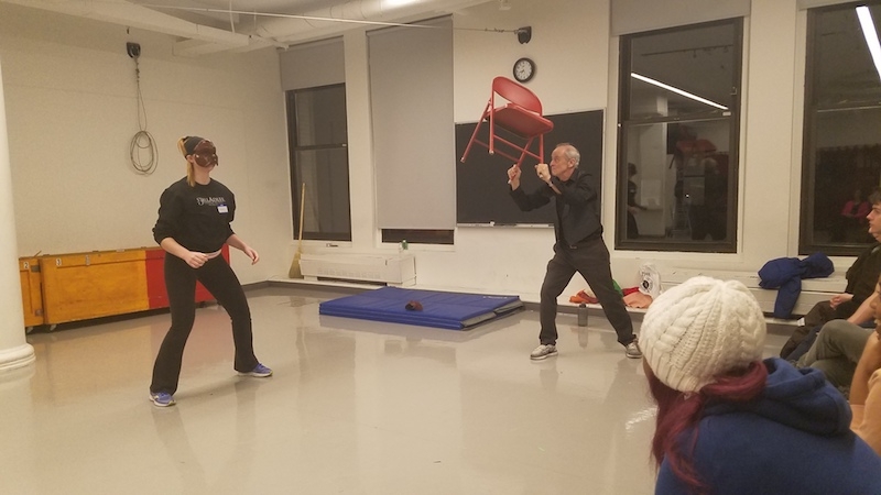 Jim Calder throwing a red chair to a student wearing a Commedia mask.